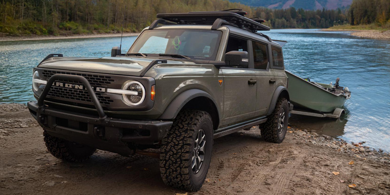 Green 2026 Ford Bronco SUV parked by a riverside in Medford, WI, towing a small boat, featuring rugged off-road tires, a front brush guard, roof rack, and a scenic forested landscape.