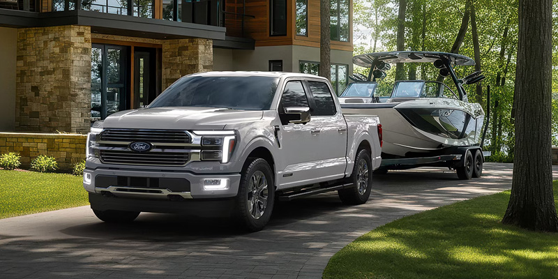 A silver Ford F-150 towing a boat on a trailer, driving along a paved driveway surrounded by trees, showcasing its towing capability and robust design.