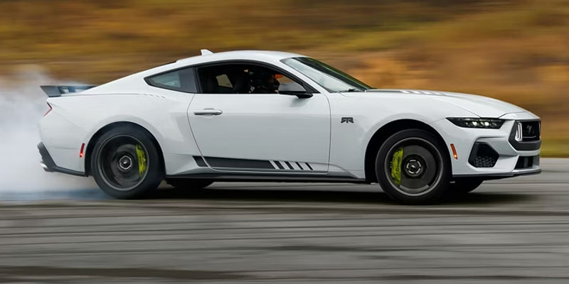 A white 2026 Ford Mustang sports car drifting on a racetrack, with smoke coming from the rear tires.