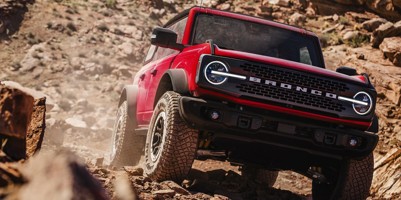 Red Ford Bronco driving over rocky terrain during an off-road adventure, showcasing its rugged design, large tires, and outdoor capability.